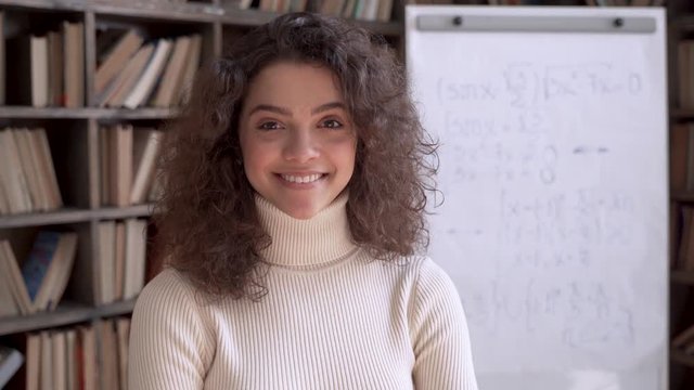 Confident smiling hispanic young woman math teacher, happy latin woman college student, professional tutor looking at camera standing arms crossed in classroom with whiteboard. Slow motion portrait.