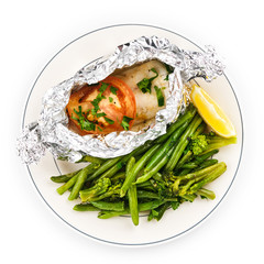 White fish fillet baked in foil with vegetables on a plate, lemon and green beans.