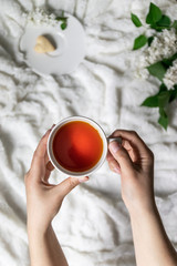 Top view of female hands holding cup with hot tea and cookies on a plate. White branches lilac on a plaid, vertical photo