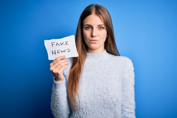 Young beautiful redhead woman holding paper with fake news message over blue background with a...