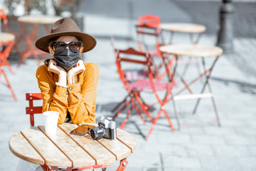 Young and sad woman in facial mask sitting on the cafe terrace alone. Concept of social distancing and new social rules after coronavirus pandemic.