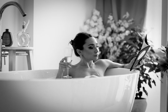 A young woman is lying in a white bath and reading a book. She holds a glass of champagne in her right hand.  Black and white version