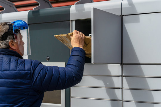 A man customer in protective face shield uses an automated self-service parcel terminal machine. Social distancing, contactless delivery, safe E-shopping, COVID-19 concept.