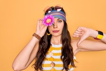 Young beautiful hippie woman with blue eyes wearing sunnglasses holding pink flower over eye with...