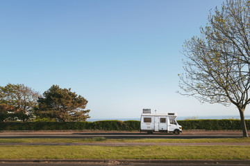 A vintage style camper van with a metal trunk attached to its roof parked on a seafront road with a...