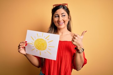 Young beautiful brunette woman holding paper with sun draw over isolated yellow background pointing and showing with thumb up to the side with happy face smiling