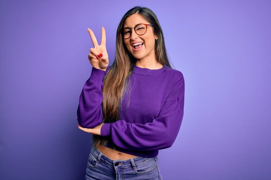 Young beautiful smart woman wearing glasses over purple isolated background smiling with happy face winking at the camera doing victory sign with fingers. Number two.
