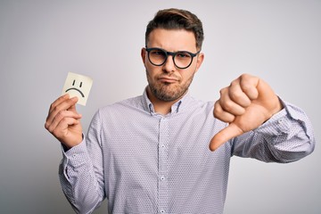 Young business man with blue eyes holding paper note with sad face as unhappy message with angry...