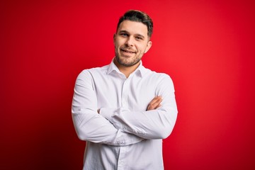 Young business man with blue eyes wearing elegant shirt standing over red isolated background happy face smiling with crossed arms looking at the camera. Positive person.