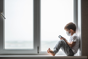 A little boy in a medical mask is sitting on the window of the house in quarantine with a phone in his hands.Prevention of coronavirus and Covid - 19