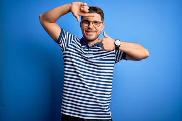 Naklejka premium Young man with blue eyes wearing glasses and casual striped t-shirt over blue background smiling making frame with hands and fingers with happy face. Creativity and photography concept.