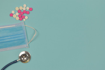 Top view of stethoscope with medical mask and pills on blue background.
