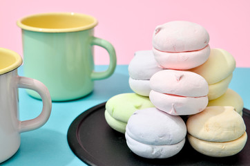 Top view of pastel colored marshmallow on black plate and two empty cups on a blue pink background. Minimalism photo for your design