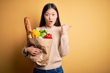 Young asian woman holding paper bag of fresh healthy groceries over yellow isolated background Surprised pointing with hand finger to the side, open mouth amazed expression.
