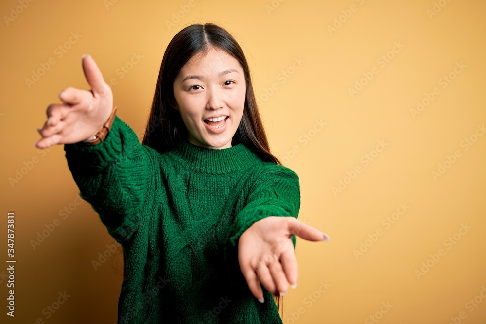 Wall mural Young beautiful asian woman wearing green winter sweater over yellow isolated background looking at the camera smiling with open arms for hug. Cheerful expression embracing happiness.