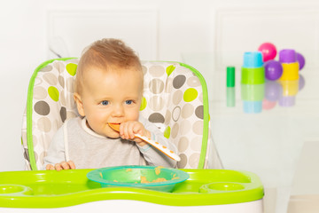 Toddler boy having thoughtful look and baby plastic spoon in mouth sit in high chair with porridge plate