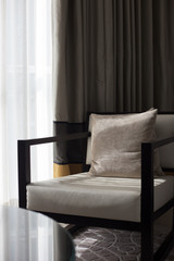 Beige armchair with pillow stands next to the table near the window with brown and white curtains, vertical orientation. Retro style