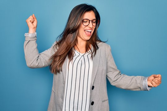 Young hispanic business woman wearing glasses standing over blue isolated background Dancing happy and cheerful, smiling moving casual and confident listening to music