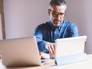 Good looking middle aged modern man having an online meeting on his laptop and tablet from home with his colleagues