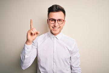Young handsome business mas wearing glasses and elegant shirt over isolated background showing and pointing up with finger number one while smiling confident and happy.