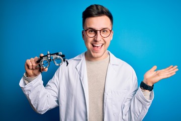 Young handsome professional optic man holding optometrical glasses over blue background very happy and excited, winner expression celebrating victory screaming with big smile and raised hands