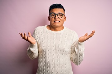 Young handsome latin man wearing white casual sweater and glasses over pink background clueless and...