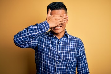 Young handsome latin man wearing casual shirt and glasses over yellow background smiling and laughing with hand on face covering eyes for surprise. Blind concept.