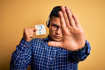 Young latin call center agent man holding question mark reminder paper using headset with open hand doing stop sign with serious and confident expression, defense gesture