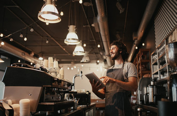 Fototapeta na wymiar Smiling caucasian man wearing apron standing behind cafe counter using digital tablet looking away