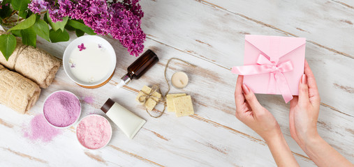 Present box in female hands. Spa and bath cosmetics with lilac flowers. Bath salt, soap, cream, oil, serum and towel rolls. Organic natural cosmetics. Beauty and relax healthcare. Gift certificate