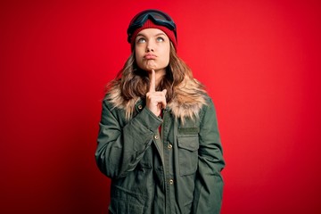 Young blonde girl wearing ski glasses and winter coat for ski weather over red background Thinking...