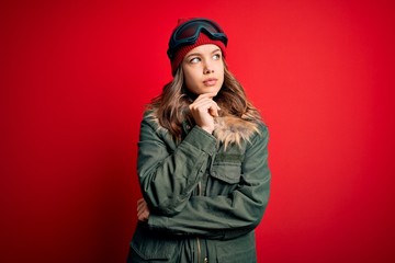 Young blonde girl wearing ski glasses and winter coat for ski weather over red background with hand on chin thinking about question, pensive expression. Smiling with thoughtful face. Doubt concept.