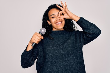 Young african american curly singer woman singing using microphone over white background with happy face smiling doing ok sign with hand on eye looking through fingers