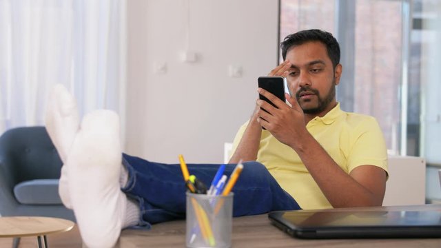 technology, remote job and people concept - stresses indian man with smartphone resting feet on table at home office