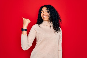 Young african american curly woman wearing casual turtleneck sweater over red background smiling with happy face looking and pointing to the side with thumb up.