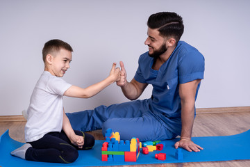 Young male doctor with beard in a blue uniform sitting on the floor next to the boy 10 years, they play educational toys