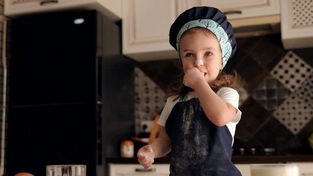 A little funny girl 2-3 years old in a chef's hat eats flour with her hands. The child indulges in the kitchen while there is no mother, eating raw dough and flour. Little girl smeared with flour.