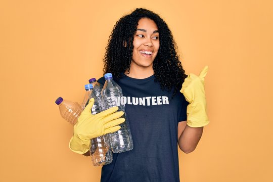 Young african american woman doing volunteering recycling holding plastic bottles pointing and showing with thumb up to the side with happy face smiling