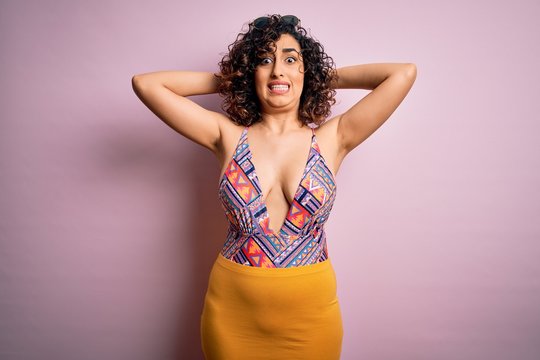 Young beautiful arab woman on vacation wearing swimsuit and sunglasses over pink background Crazy and scared with hands on head, afraid and surprised of shock with open mouth