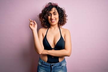 Young beautiful arab woman on vacation wearing swimsuit and sunglasses over pink background with a big smile on face, pointing with hand and finger to the side looking at the camera.