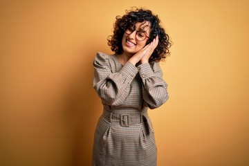 Fototapeta premium Beautiful arab business woman wearing dress and glasses standing over yellow background sleeping tired dreaming and posing with hands together while smiling with closed eyes.