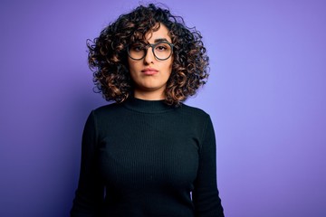 Young beautiful curly arab woman wearing casual sweater and glasses over purple background with serious expression on face. Simple and natural looking at the camera.