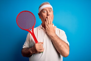 Middle age hoary sportsman playing tennis using racket over isolated blue background cover mouth with hand shocked with shame for mistake, expression of fear, scared in silence, secret concept