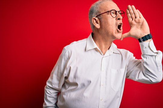 Middle age handsome hoary man wearing casual shirt and glasses over red background shouting and screaming loud to side with hand on mouth. Communication concept.