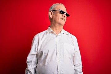 Middle age hoary man wearing funny sunglasses over isolated red background looking away to side with smile on face, natural expression. Laughing confident.