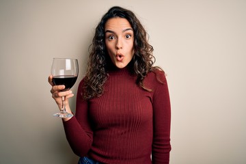 Young beautiful woman with curly hair drinking glass of red wine over white background scared in...