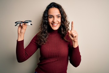 Young beautiful woman with curly hair holding glasses over isolated white background surprised with an idea or question pointing finger with happy face, number one