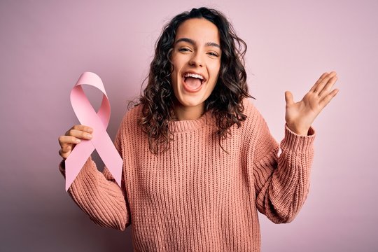 Beautiful woman with curly hair holding pink cancer ribbon symbol over isolated background very happy and excited, winner expression celebrating victory screaming with big smile and raised hands
