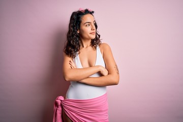 Beautiful woman with curly hair on vacation wearing white swimsuit over pink background looking to the side with arms crossed convinced and confident