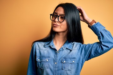 Young beautiful chinese woman wearing casual denim shirt over isolated yellow background confuse and wondering about question. Uncertain with doubt, thinking with hand on head. Pensive concept.